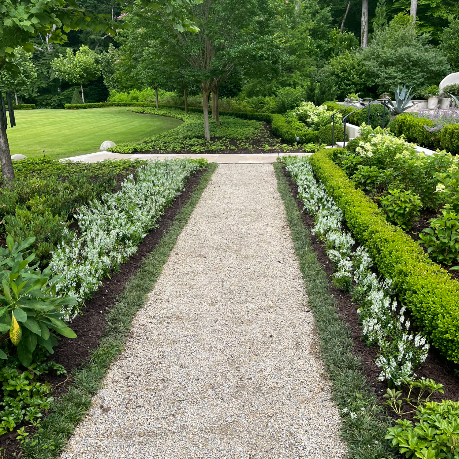 Beautifully landscaped gravel garden pathway in Atlanta lined with white flowering plants, manicured boxwoods, lush greenery, and mature trees, expertly designed by Gibbs Landscape Company.