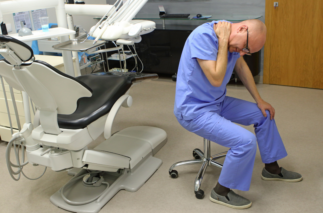 Dentist in blue scrubs sitting on a stool holding his neck in pain next to a dental chair in clinic.