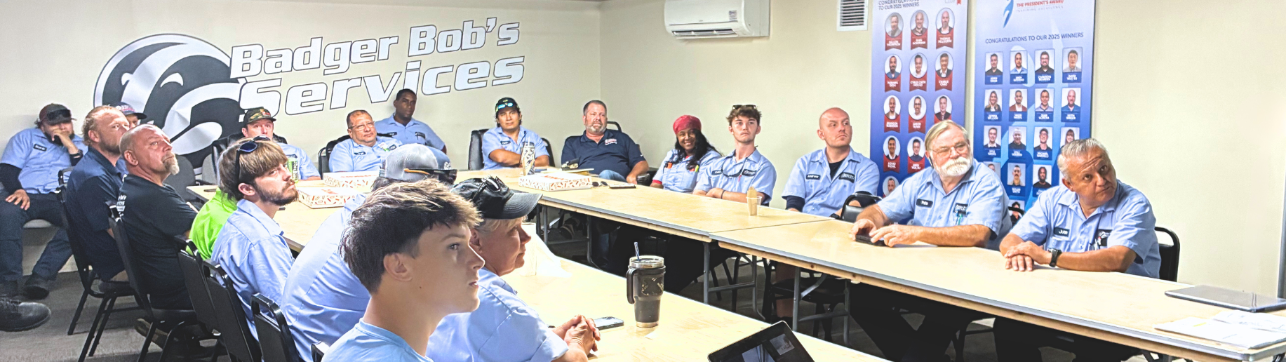 Employees in blue uniforms attend a meeting in a conference room with Badger Bob's Services logo on the wall.