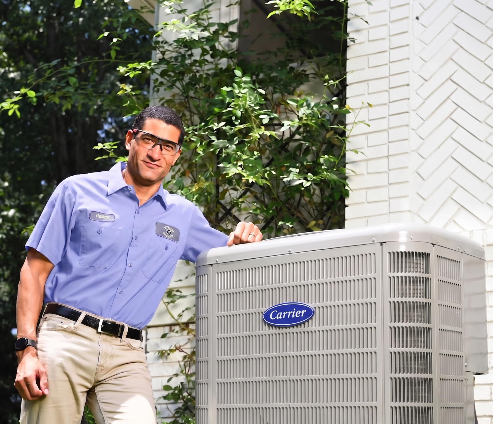 Technician in blue shirt and safety glasses standing next to a Carrier air conditioning unit outdoors.
