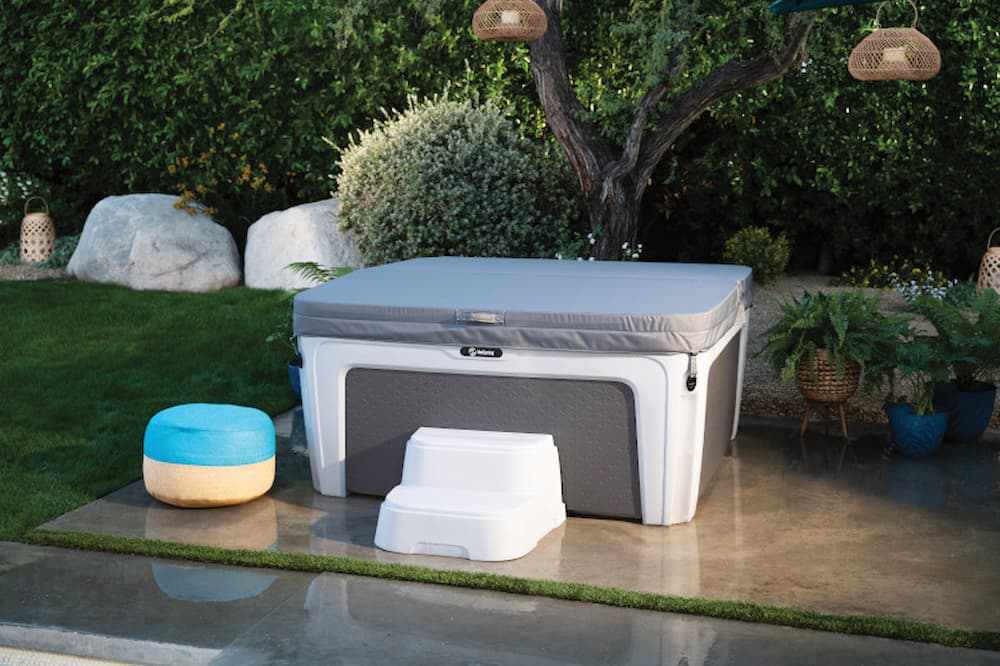 Covered gray and white hot tub with steps and a round blue cushion in a landscaped backyard.