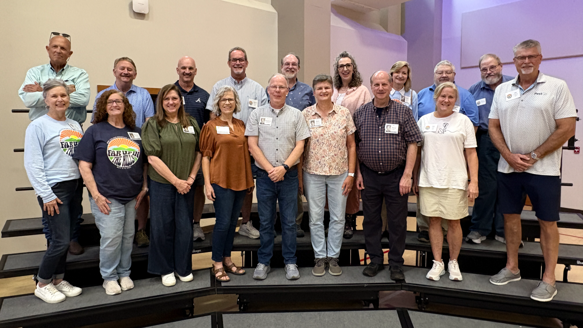 Group photo of 18 adults standing on risers indoors in casual clothing with name tags.