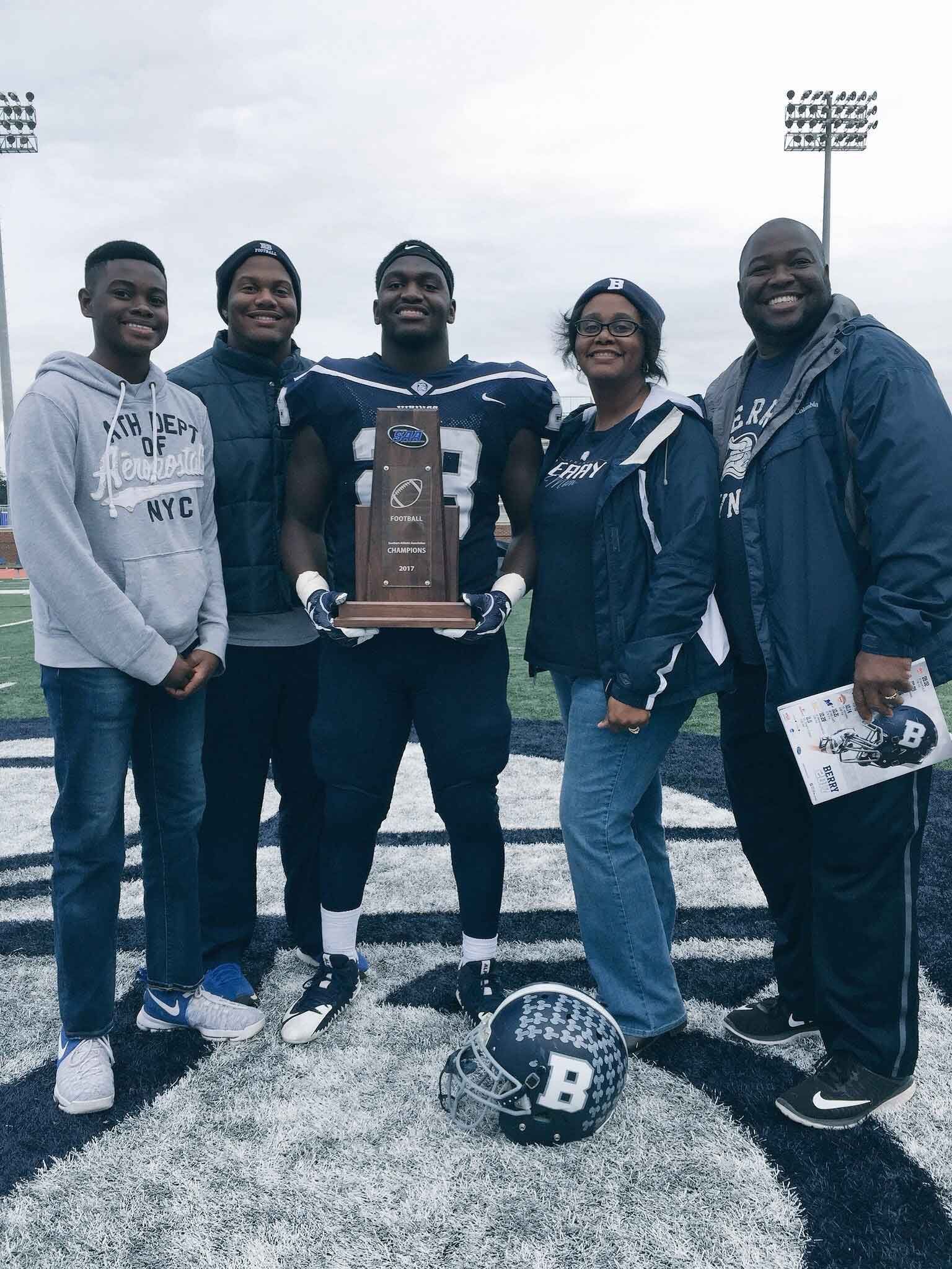 A family surrounds their son who holds up a football championship trophy.