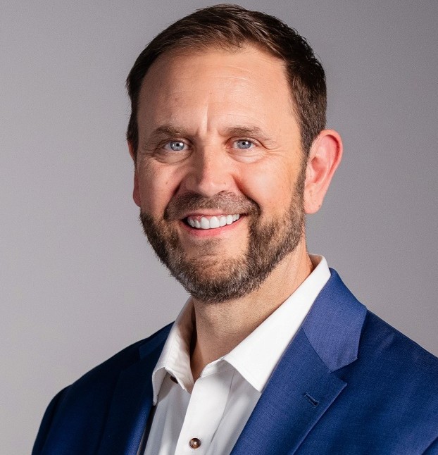 Smiling bearded man with blue eyes wearing a blue blazer and white shirt against a gray background.