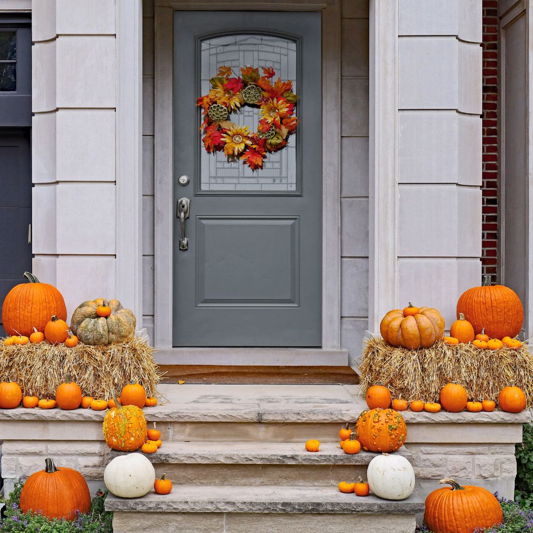 Front porch decorated with various pumpkins, hay bales, and an autumn leaf wreath on a gray door for fall.