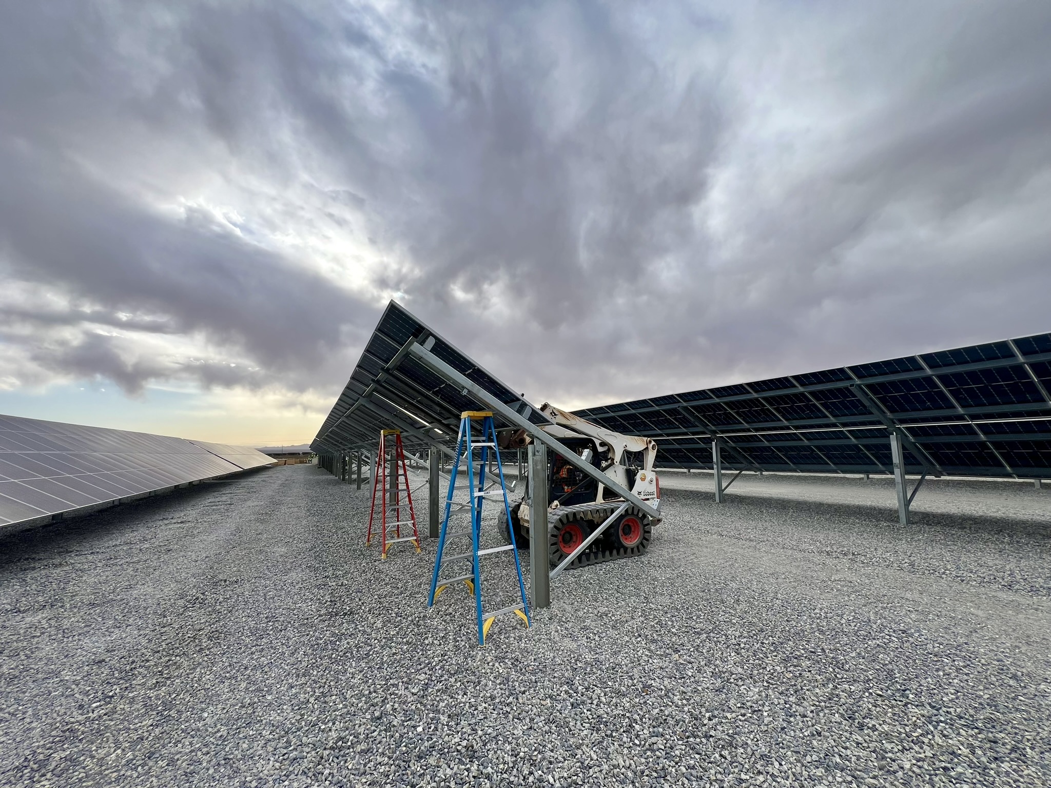 Solar panels being installed on a gravel field with ladders and a small construction vehicle under cloudy sky.