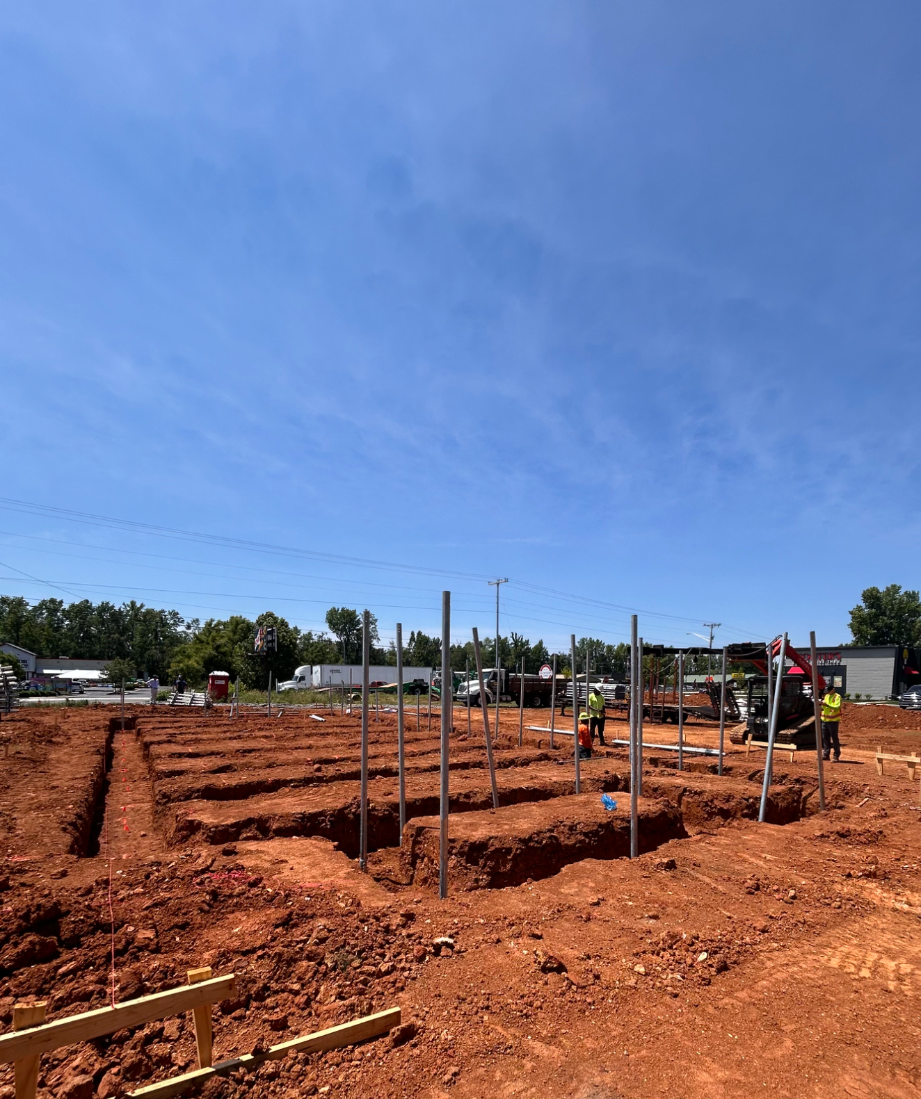 Construction site with trenches and Cantsink helical pilesunder a clear blue sky and workers in high-visibility clothing.