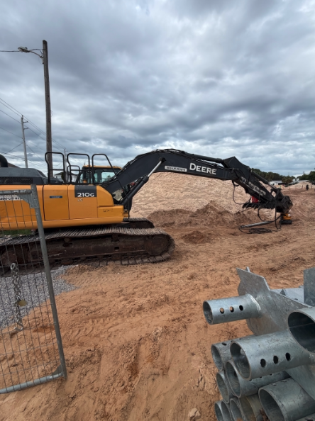 Excavator installing helical piles on sandy construction site under cloudy sky.