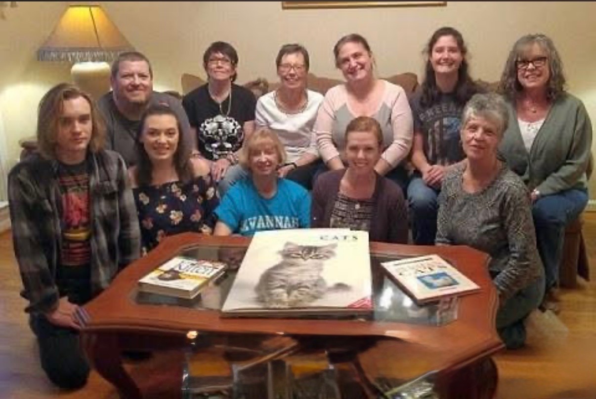 Group of twelve people smiling around a coffee table with books, including a cat book, in a cozy living room.