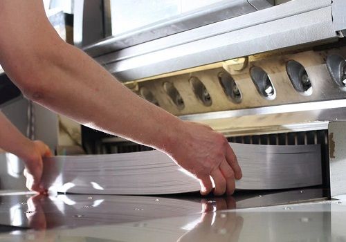 A printing plant worker placing a thick stack of paper sheets into a large paper shear