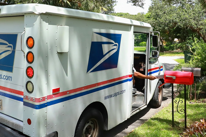 A mail carrier reaching out of his truck to put mail in a red mailbox