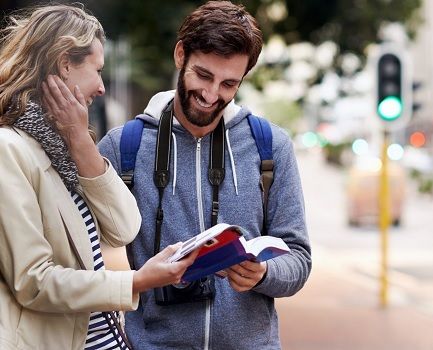 A female tourist and a male tourist reading a travel guide while standing near a busy street rourist