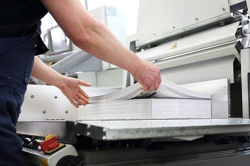 A worker in a printing plant gathering some printed book pages from a large stack.