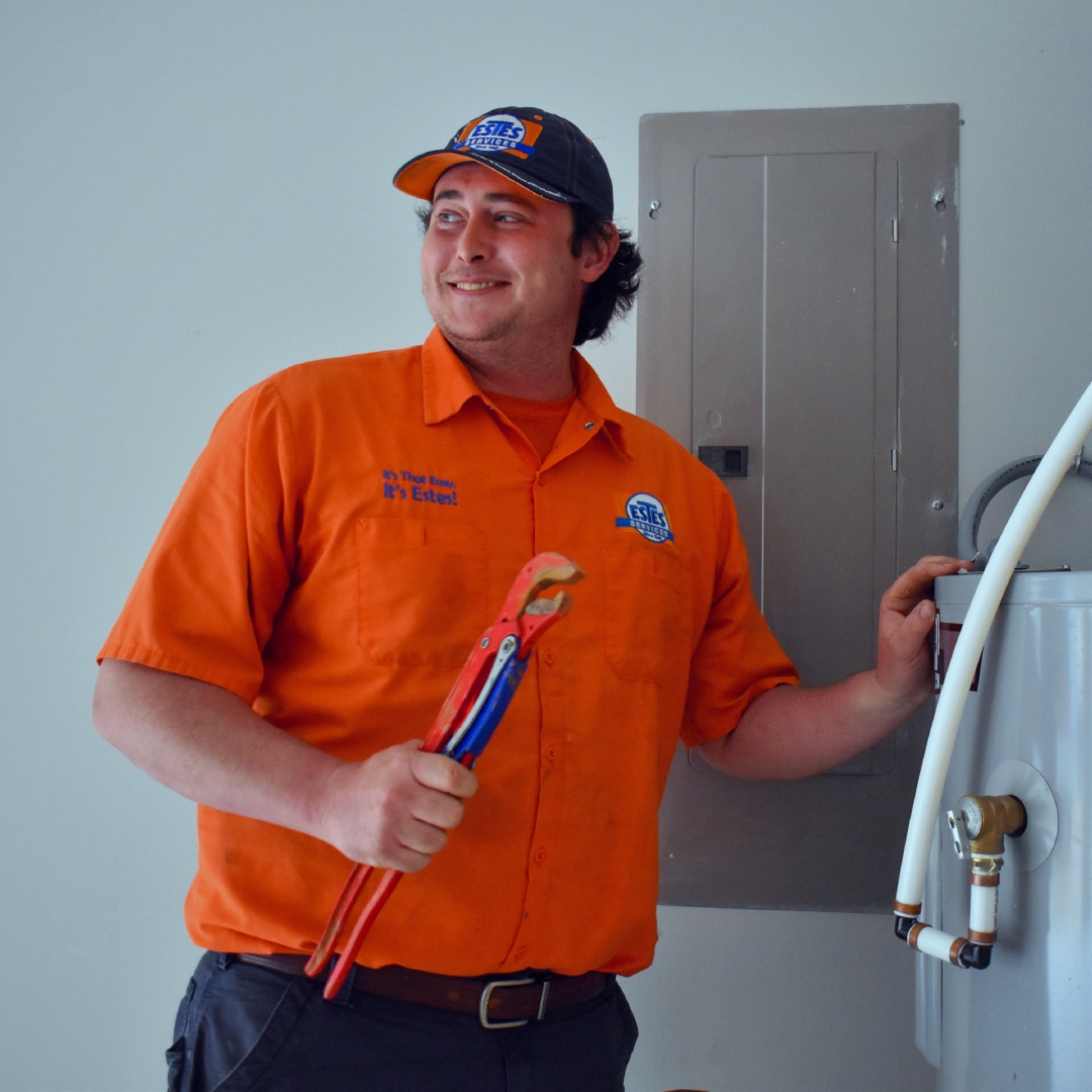 Estes Smiling plumber in orange uniform holding pipe wrench next to water heater and electrical panel indoors.