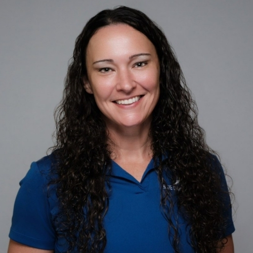 Smiling woman with long curly dark hair wearing a blue polo shirt against a plain gray background