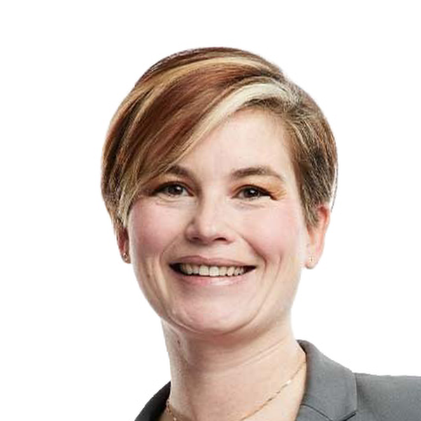 Smiling woman with short brown and blonde hair wearing a gray blazer and gold necklace on white background