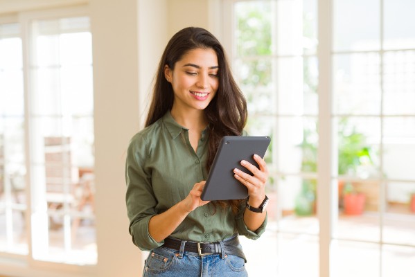 Young woman smiling and using a tablet in a bright room with large windows and plants in the background