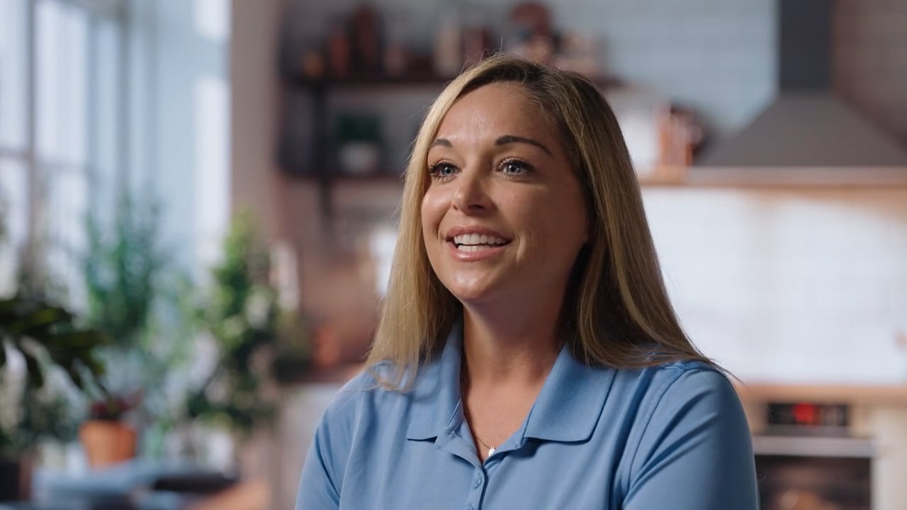Smiling woman with long blonde hair wearing a blue polo shirt in a cozy, well-lit kitchen setting.