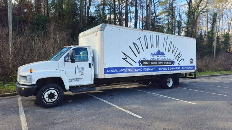 White Midtown Moving truck parked in an empty lot near trees with company logo and services on the side.