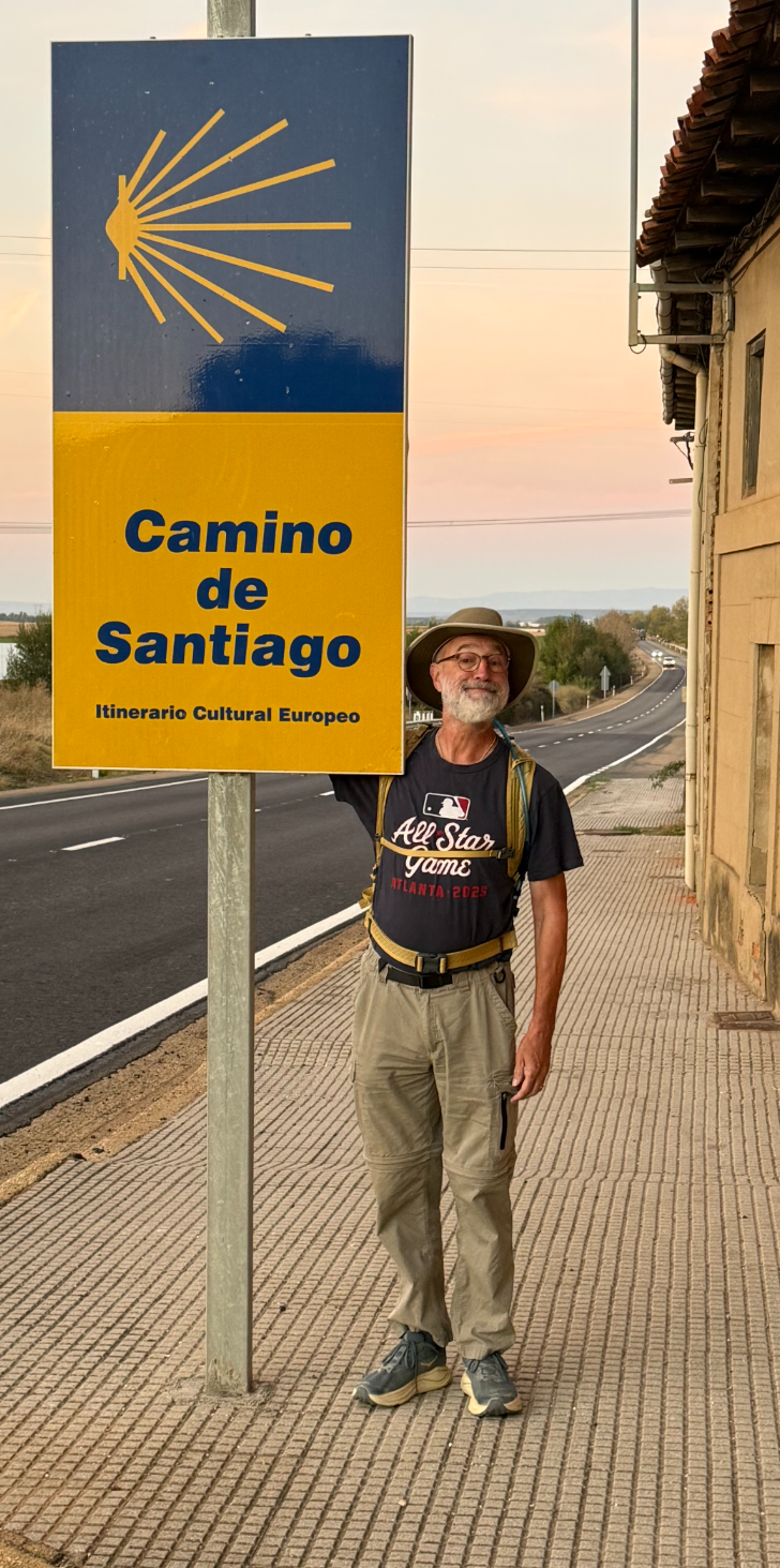 Man standing beside a Camino de Santiago road sign on a rural sidewalk with an orange building and empty road at sunset.