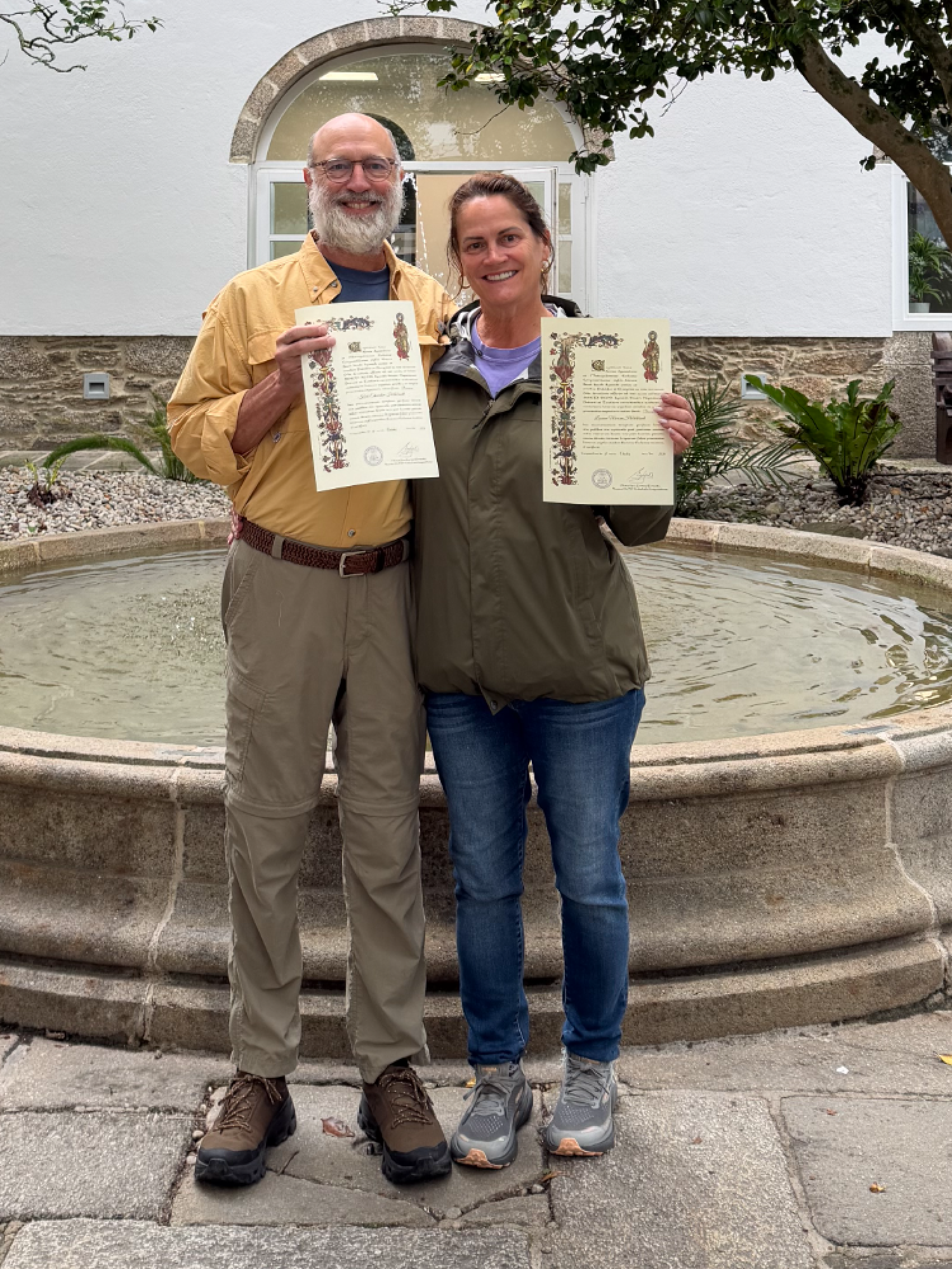 Smiling couple standing by a stone fountain holding official certificates in a courtyard with trees and white building.