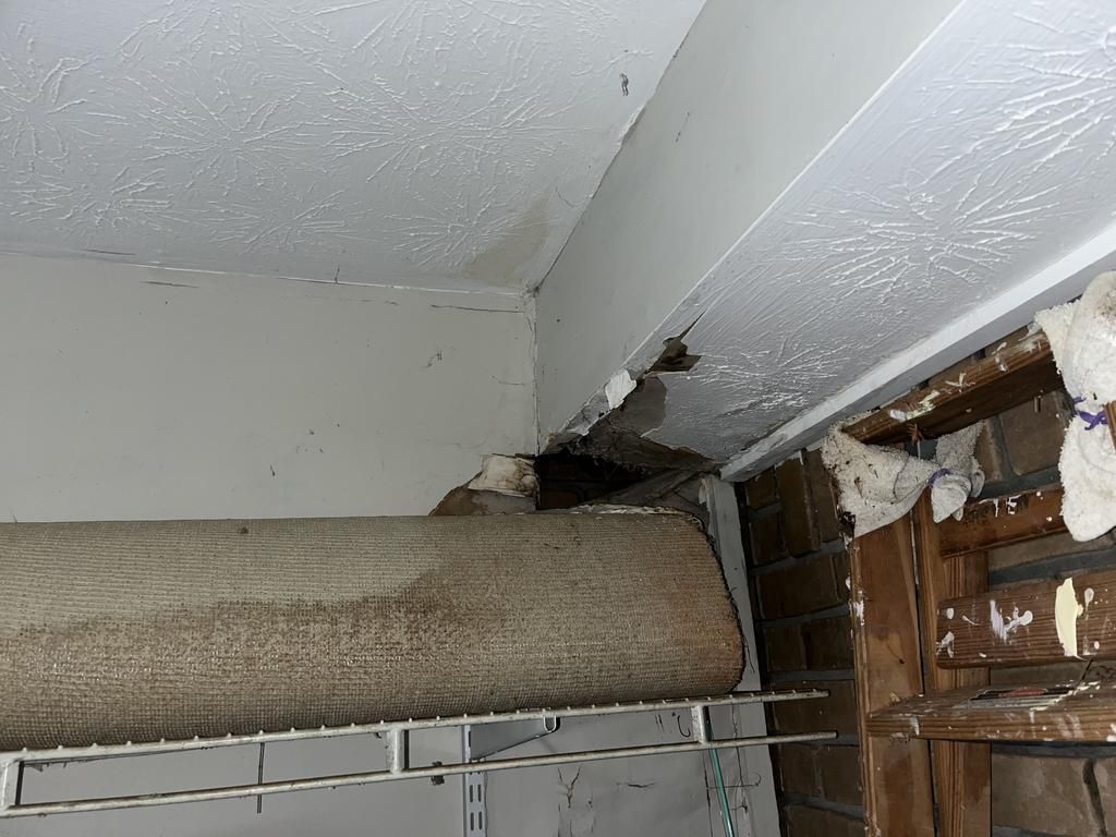 Damaged interior ceiling and wall corner with cracks, peeling paint, and broken drywall exposing wooden framing and insulation, with a rolled-up carpet on a shelf below in Marietta