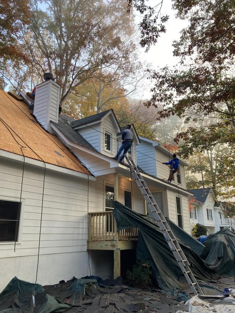 Residential roof renovation with Nelson Exterior employees replacing shingles and repairing exposed wooden sheathing, tarps covering parts of the house and ground, and autumn trees surrounding the two-story white home in Marietta