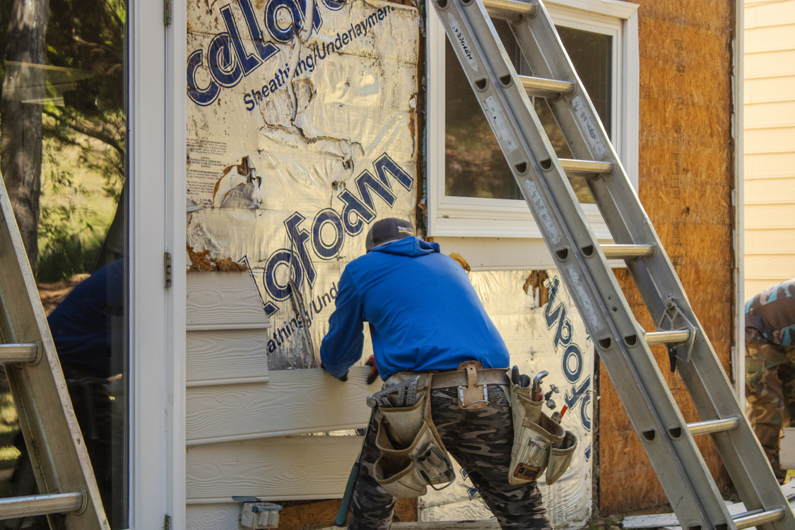 Worker installing exterior siding on a house with ladder and construction tools during renovation.