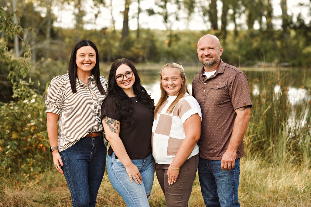 Smiling family of four standing outdoors near a lake with trees and grass in the background.