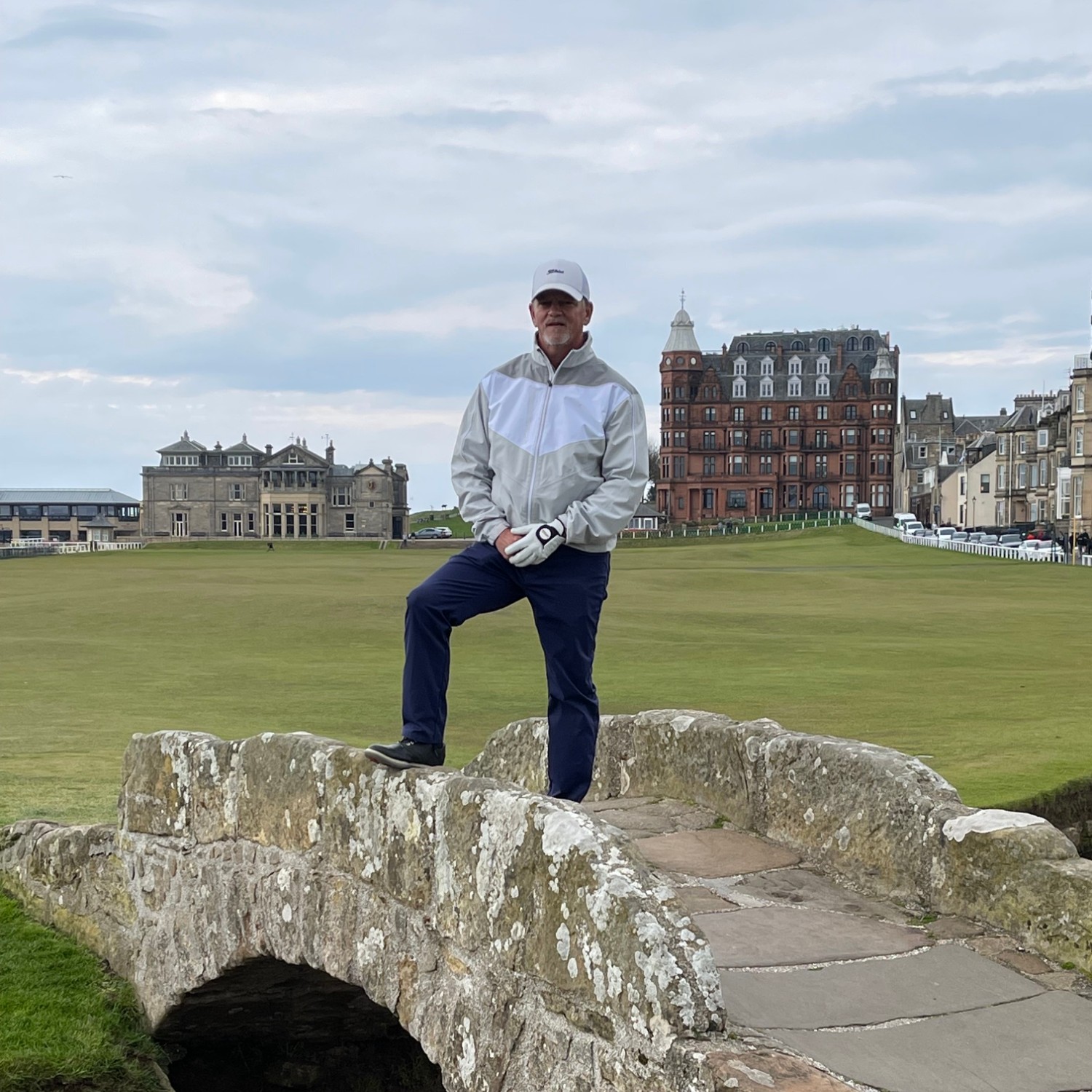 Man in golf attire standing on historic stone bridge over a small stream with golf course and buildings in background.