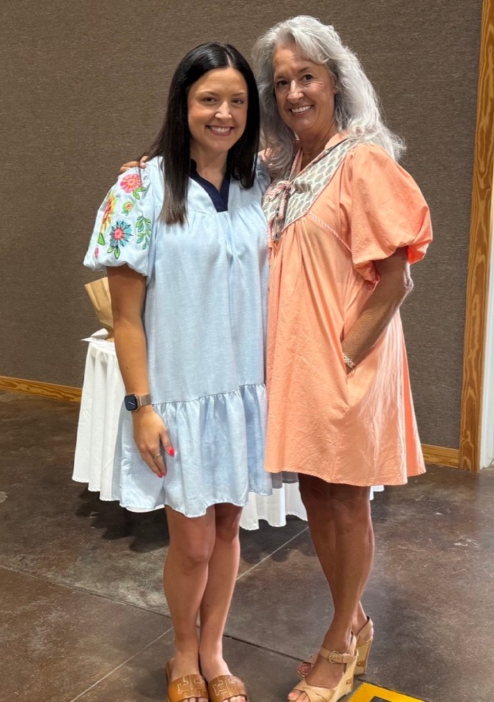 Two women smiling indoors wearing pastel short dresses with floral embroidery and sandals.