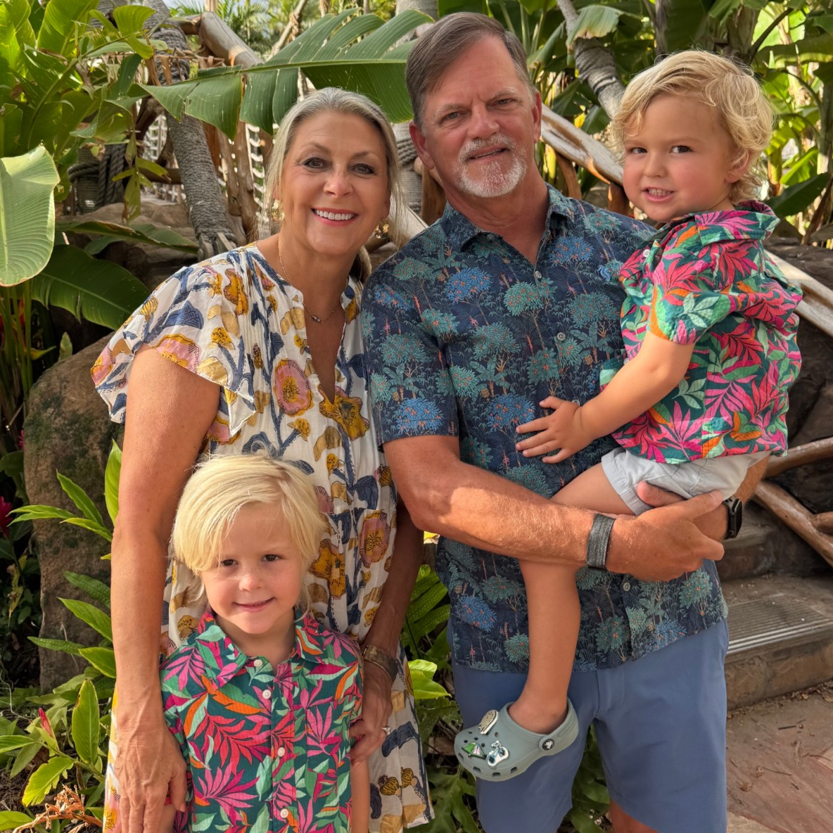 Happy family of four posing outdoors in tropical garden with vibrant green foliage and sunny weather.