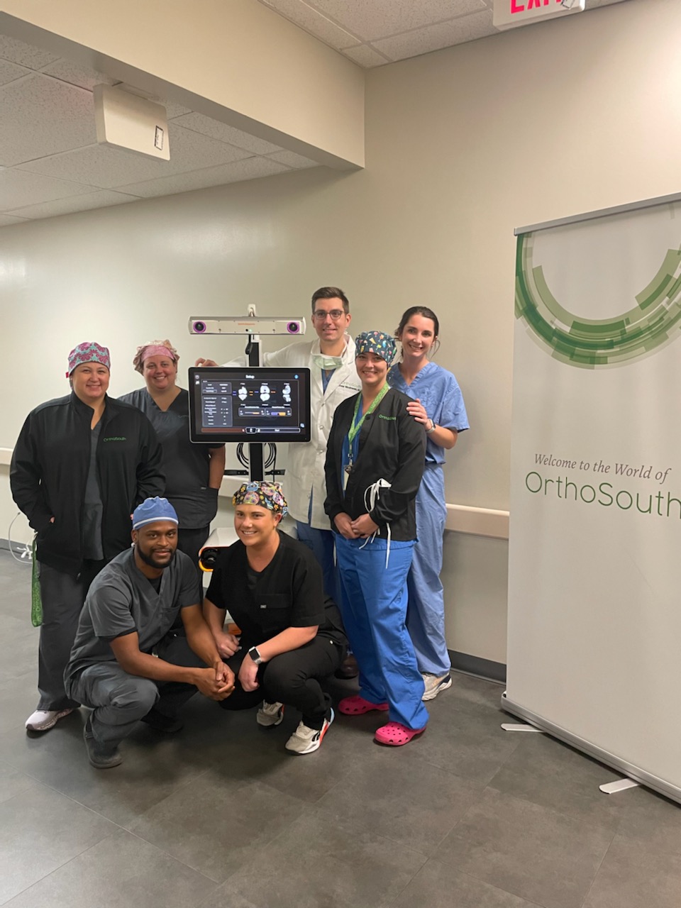 Group of medical professionals in scrubs and lab coats posing with surgical equipment next to OrthoSouth banner.