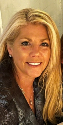 Smiling blonde woman with long hair wearing a dark patterned jacket and necklace in a warm indoor setting