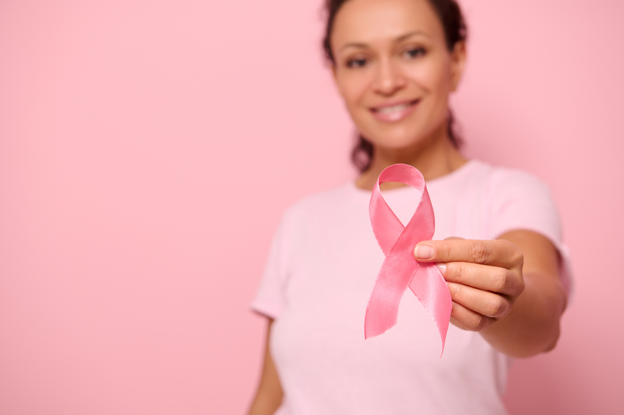 Smiling woman in pink shirt holding a pink breast cancer awareness ribbon with a pink background.