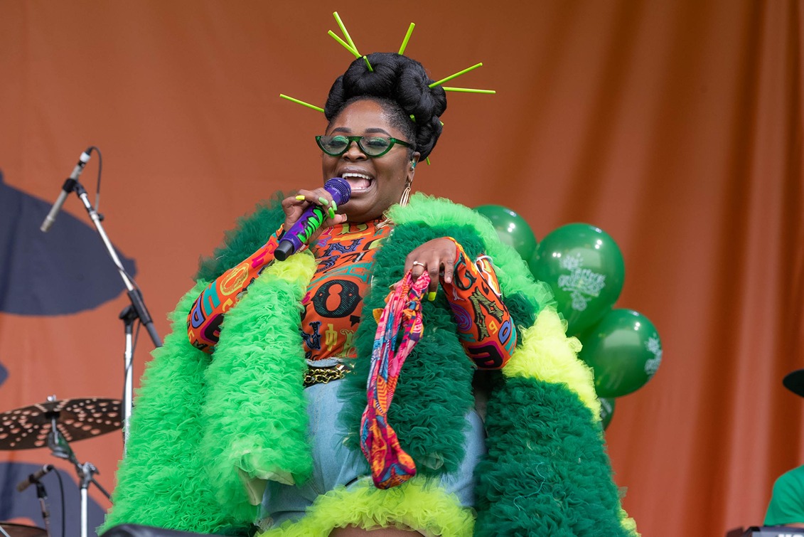 Singer in vibrant green and orange outfit with unique hair accessories performs on stage with microphone.
