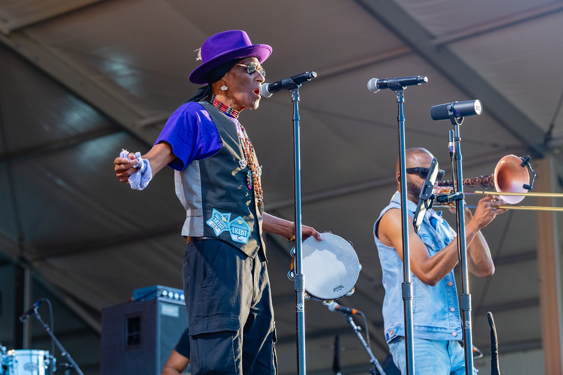 Musician in purple hat sings into microphone playing tambourine while another plays trombone at outdoor concert.