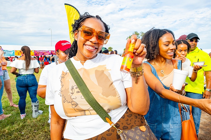 Smiling woman wearing sunglasses holds hot sauce bottle at outdoor festival with people and booths in background