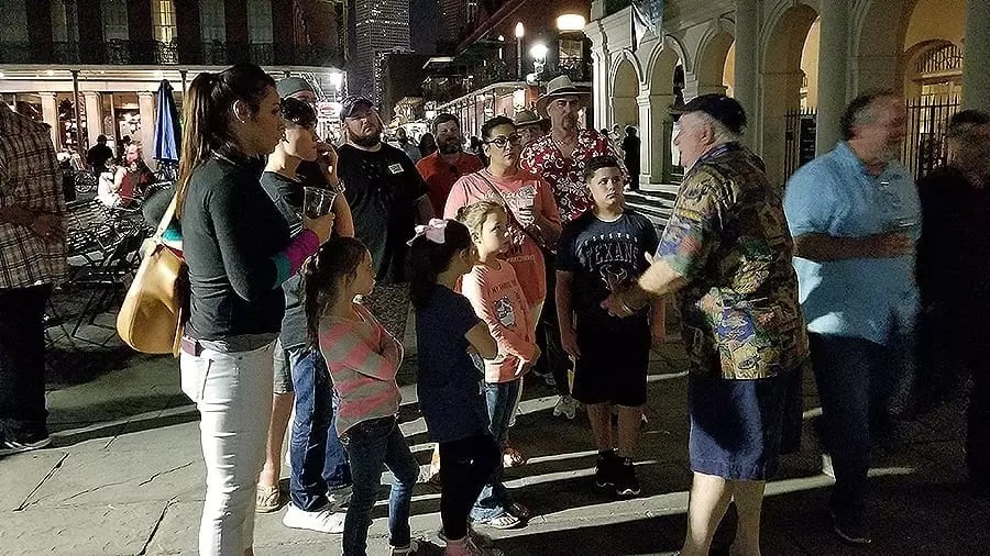 Group of people, including children and adults, listening to a man speaking outdoors at night in a historic area.