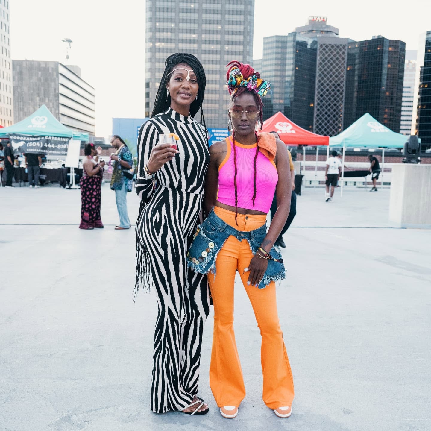 Two stylish women in bold outfits posing outdoors with city buildings and tents in the background