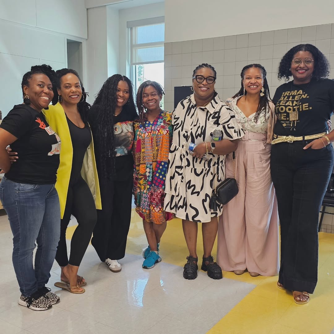 Group of seven diverse women standing together smiling in a bright indoor setting with casual and colorful outfits