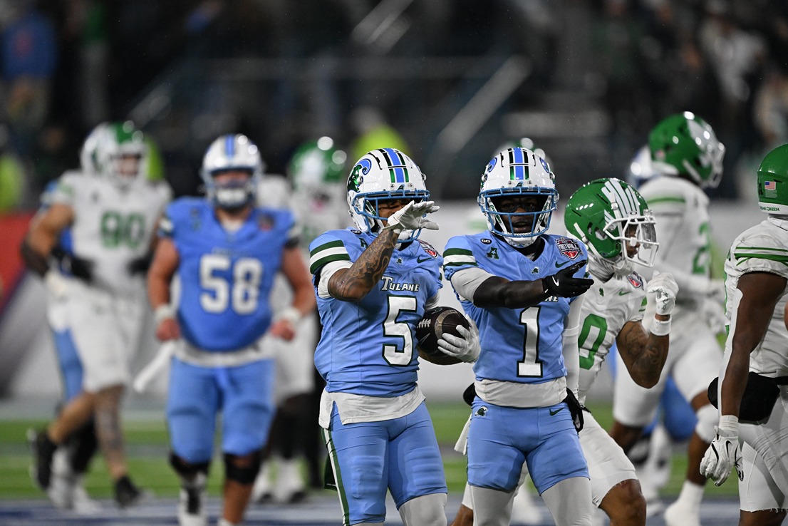 Two Tulane football players in blue uniforms celebrate with one holding the ball during a game against a team in white and green.