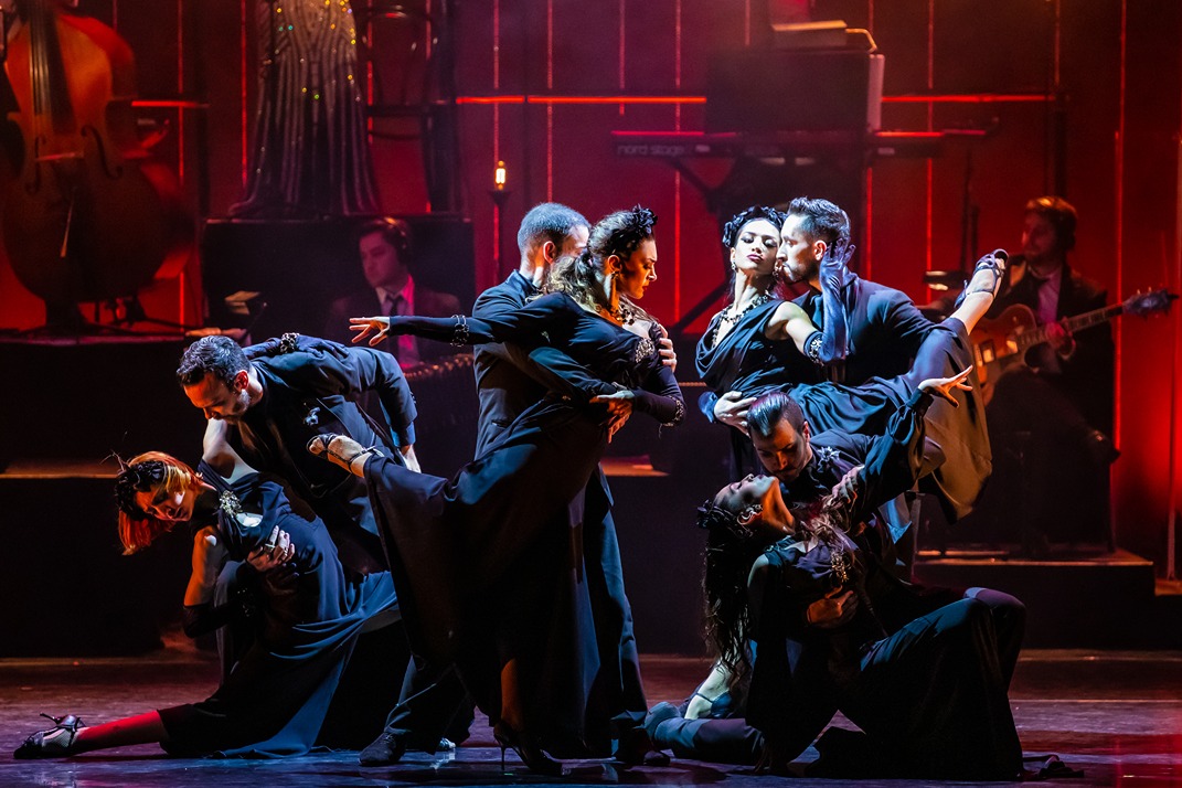 Eight dancers in elegant black outfits performing a dramatic tango on stage with warm red lighting and live musicians.