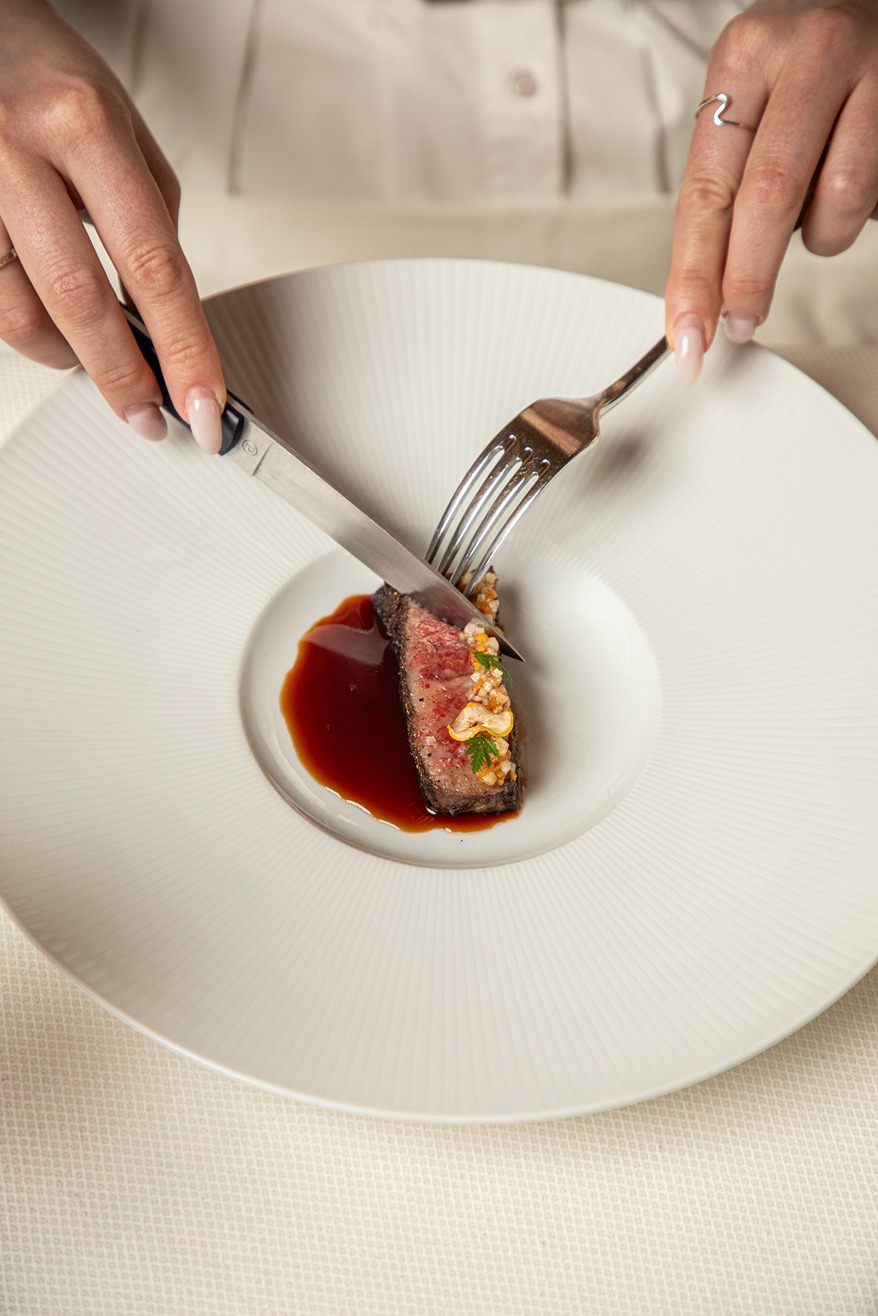 Hands cutting a small gourmet steak on a white plate with sauce and garnish in a fine dining setting