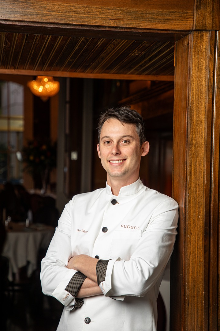 Smiling male chef in white uniform standing with arms crossed inside restaurant near wooden door.