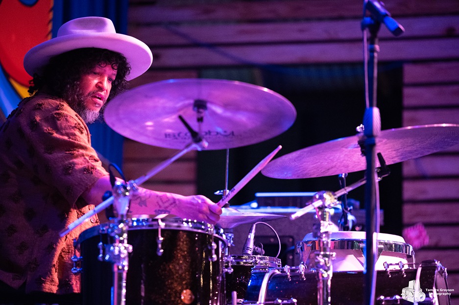 Drummer playing a drum set on stage under purple lighting wearing a hat and patterned shirt