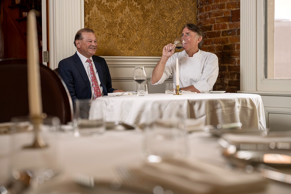 Two men at a formal dining table in an elegant restaurant, one tasting wine, the other smiling in a suit.
