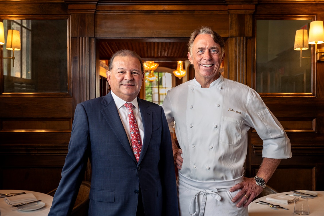 Two men smiling in a restaurant setting, one in a suit and red patterned tie, the other in a white chef's jacket.