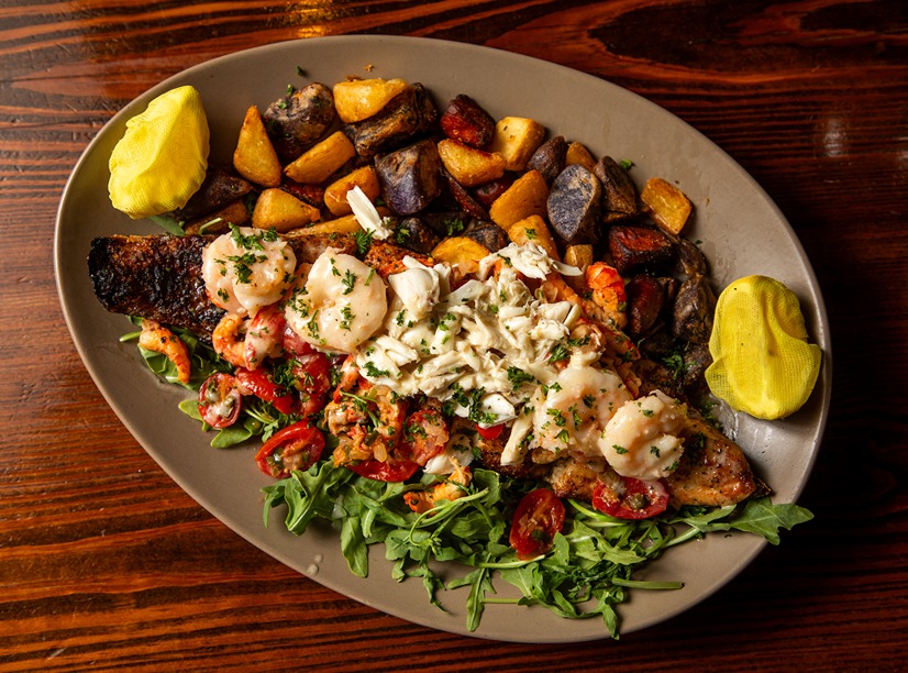 Grilled seafood platter with shrimp, crab, roasted potatoes, arugula, cherry tomatoes, and lemon wedges on a plate.
