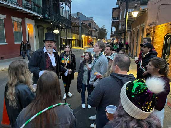 Group on historic street listening to man in top hat during dusk city tour gathering.